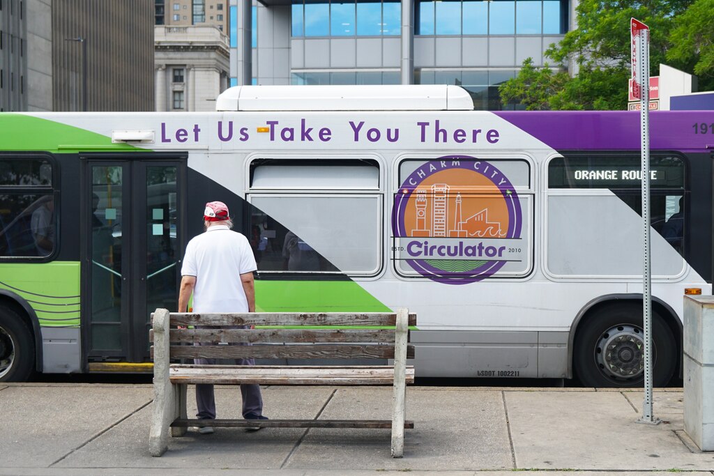 A green, white and purple bus stops at a bus stop in front of a man standing by a bench.