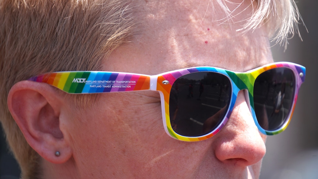 A very close up image of the face of a woman with short blonde hair wearing rainbow-colored sunglasses.