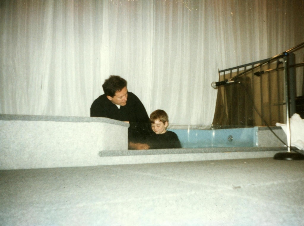 Photograph of adult white man dressed in black leaning over white boy, also in black, in a baptismal tub. There are white curtains in the background, white tile floor in the foreground, and a microphone in a stand on the right.