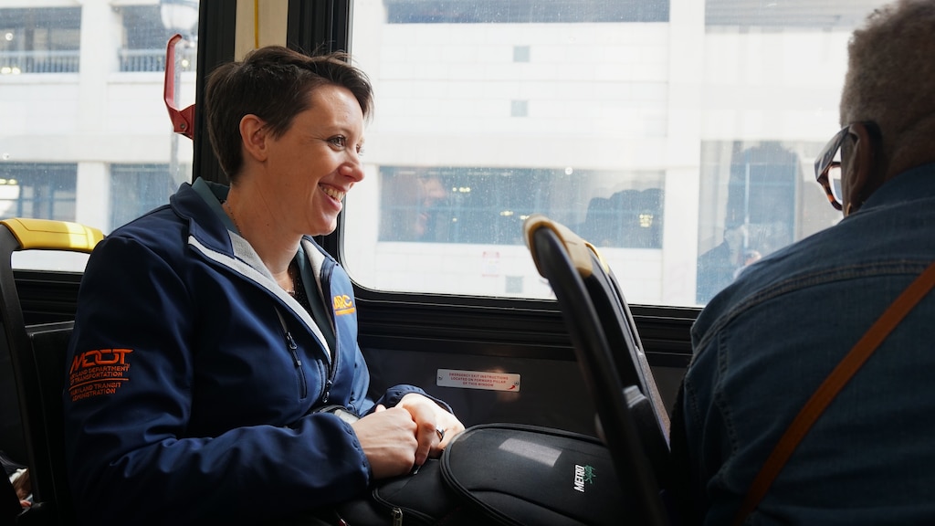 A woman with short brown hair sits in a bus seat and talks with the woman sitting in front of her.