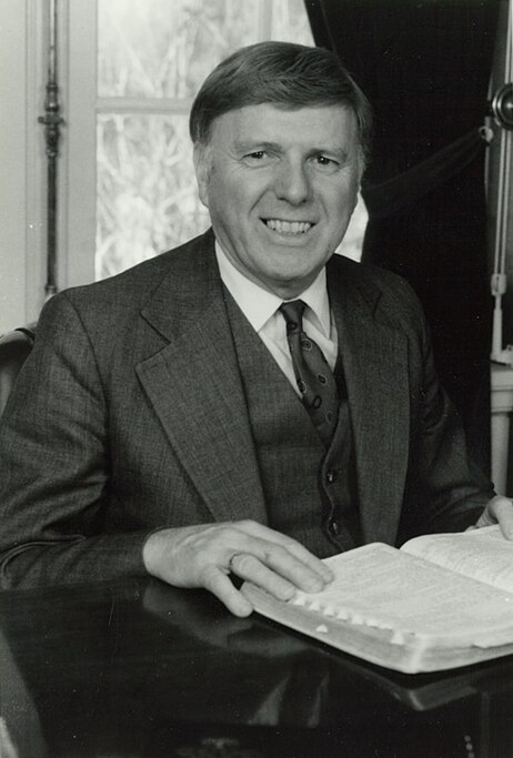 Black and white photograph of middle aged white man in three piece suit and striped tie, smiling at camera, with his hand on an open Bible in front of him on the table