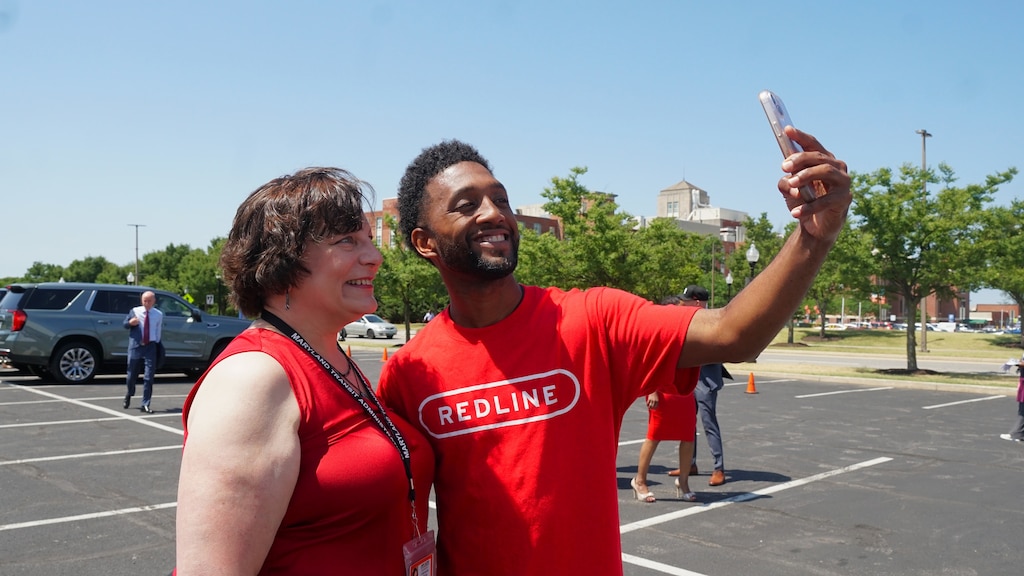 A man wearing a red t-shirt that says "REDLINE" smiles and takes a selfie with a woman in a parking lot.