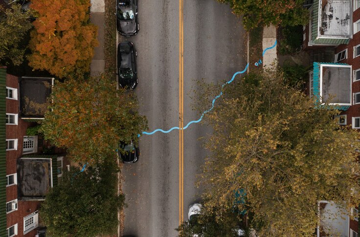 A bird's eye view of a city street, lined with trees and cars, with a blue thermoplastic line squiggling across the road as part of an art installation.