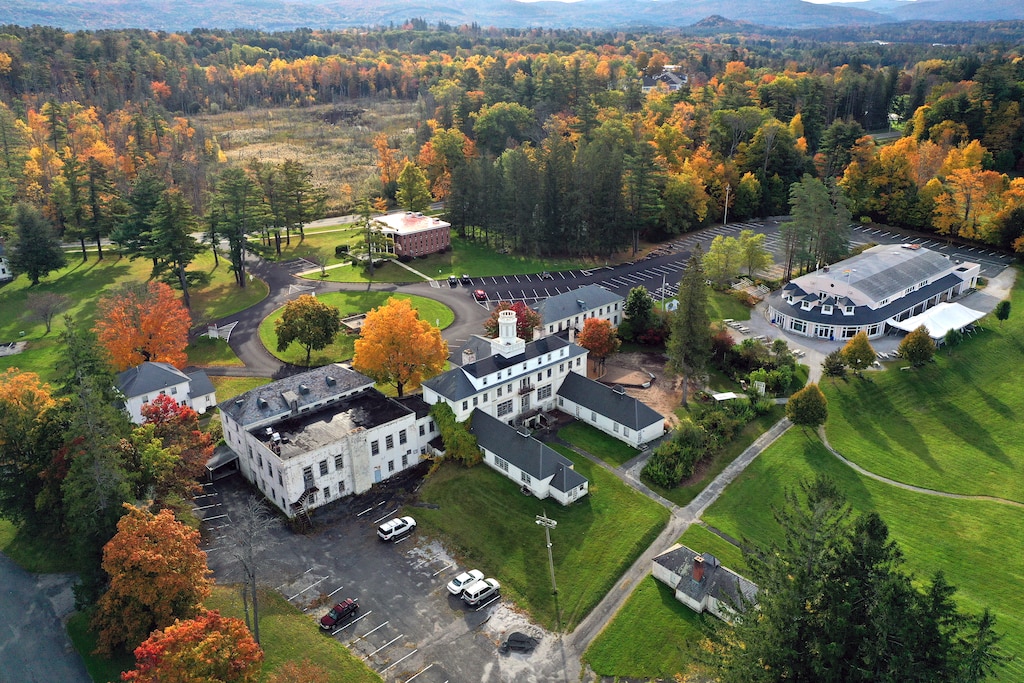 Aerial photograph of campus of several multi-story white buildings with gray roofs surrounded by green lawns and autumnal trees, with hills in the background.