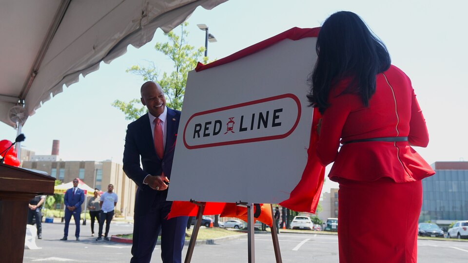 A man in a suit and tie and a woman in a red suit stand next to a poster board that reads "RED LINE" with a train symbol in between the two words.