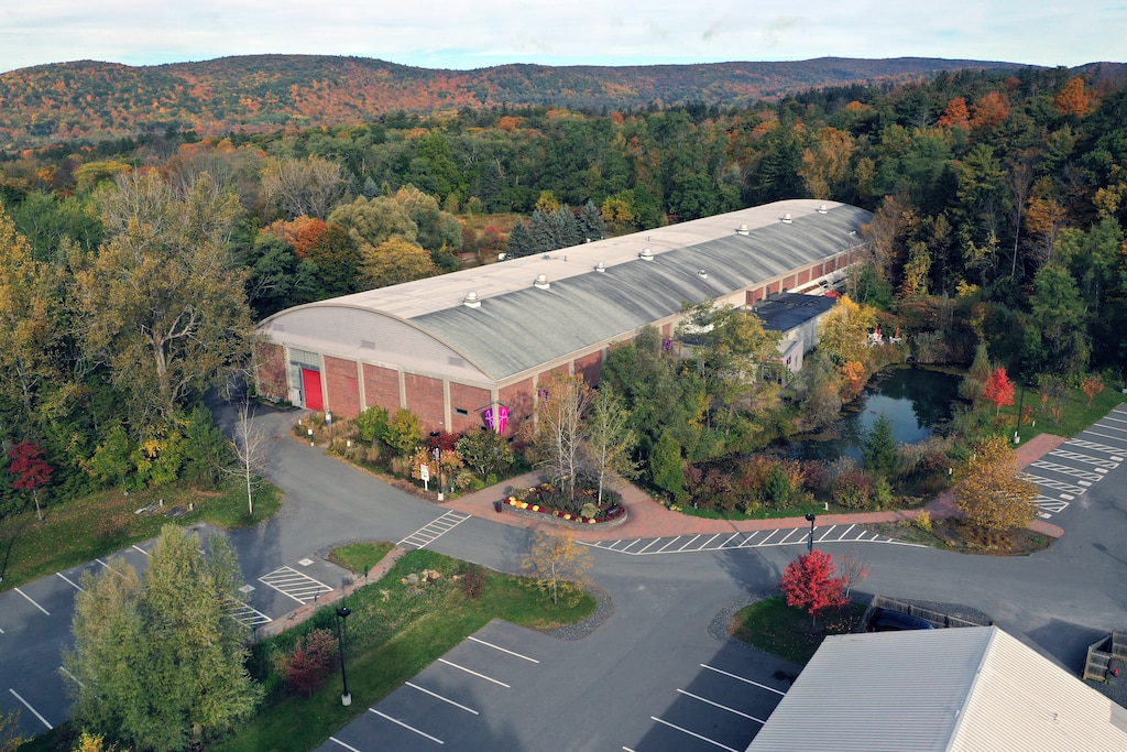Aerial photo of long rectangular building with arched roof nestled among autumnal trees and hills in the background, and parking lot in front.