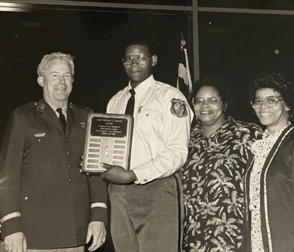 Four people stand posing for a picture as a young man, second from the left, accepts an award.