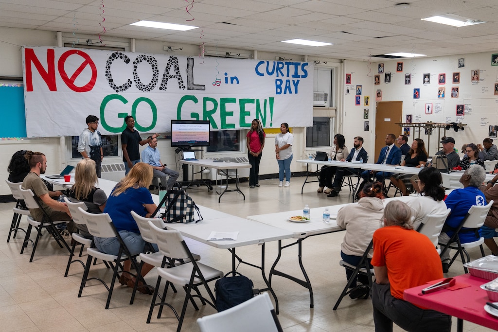 A wide shot of the Curtis Bay community meeting. A large hand-drawn banner reading "No coal in Curtis Bay, go green!" hands in the background.