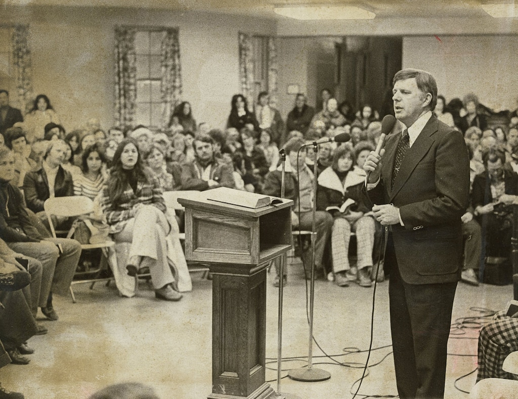 Black and white photogaph of middle aged white man standing in foreground, speaking into microphone, with crowd of seated people in background. Next to man is a podium with open book on it.