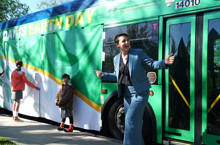 A woman in a blue suit stands in front of a green bus. Two kids stand in front of green and white panels covering the side of the bus.